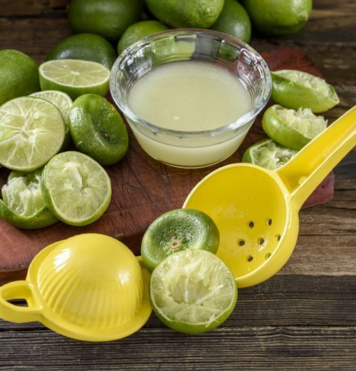 A bunch of limes and a bowl of juice on a wooden table