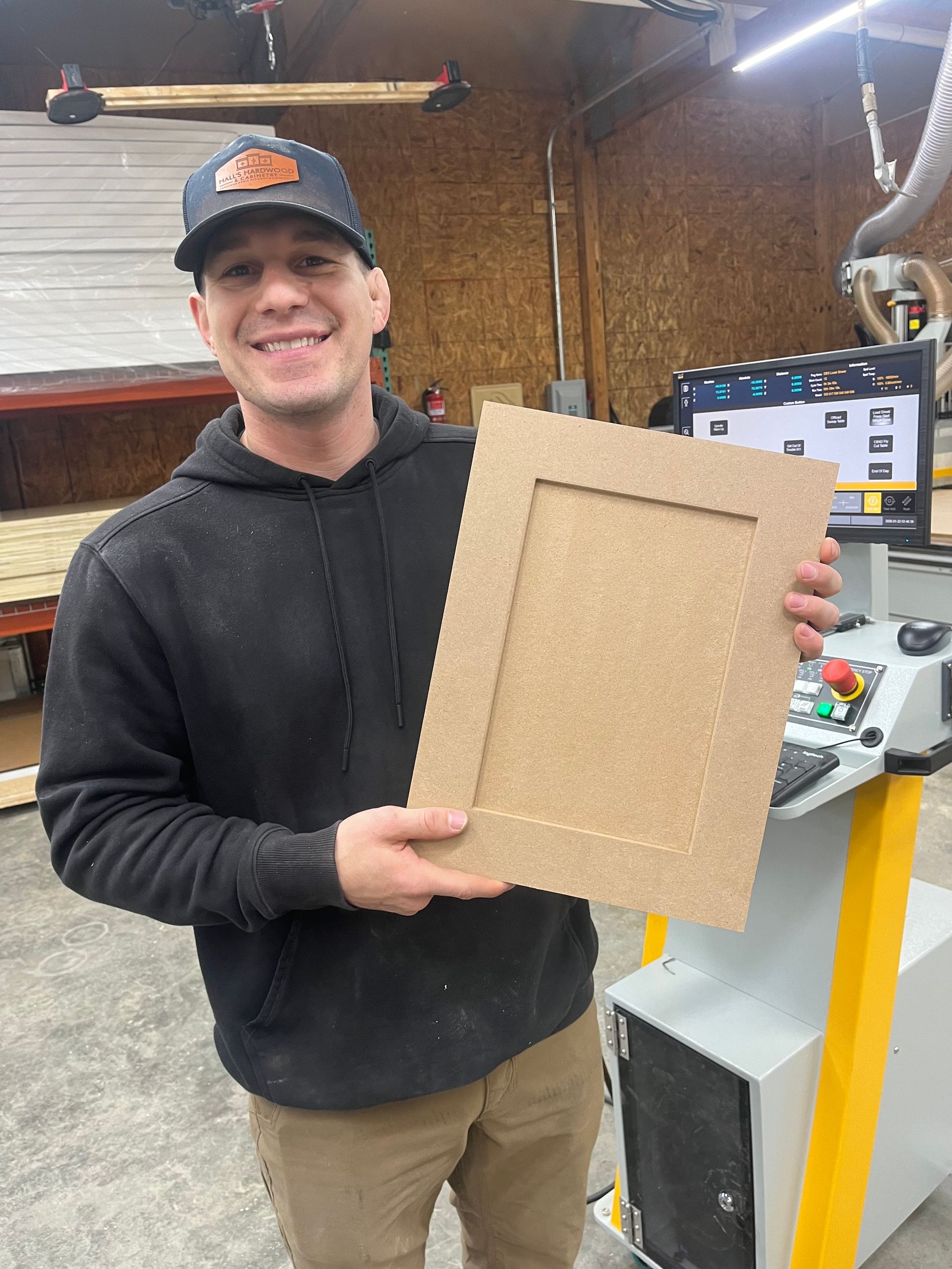 Man holding wooden cabinet door in a workshop, smiling. Computer and machinery in background.
