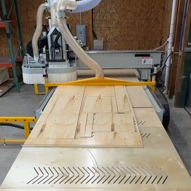 A CNC machine cutting plywood; workshop setting. Light wood material with cut patterns. Yellow safety bar and dust extraction hose visible.