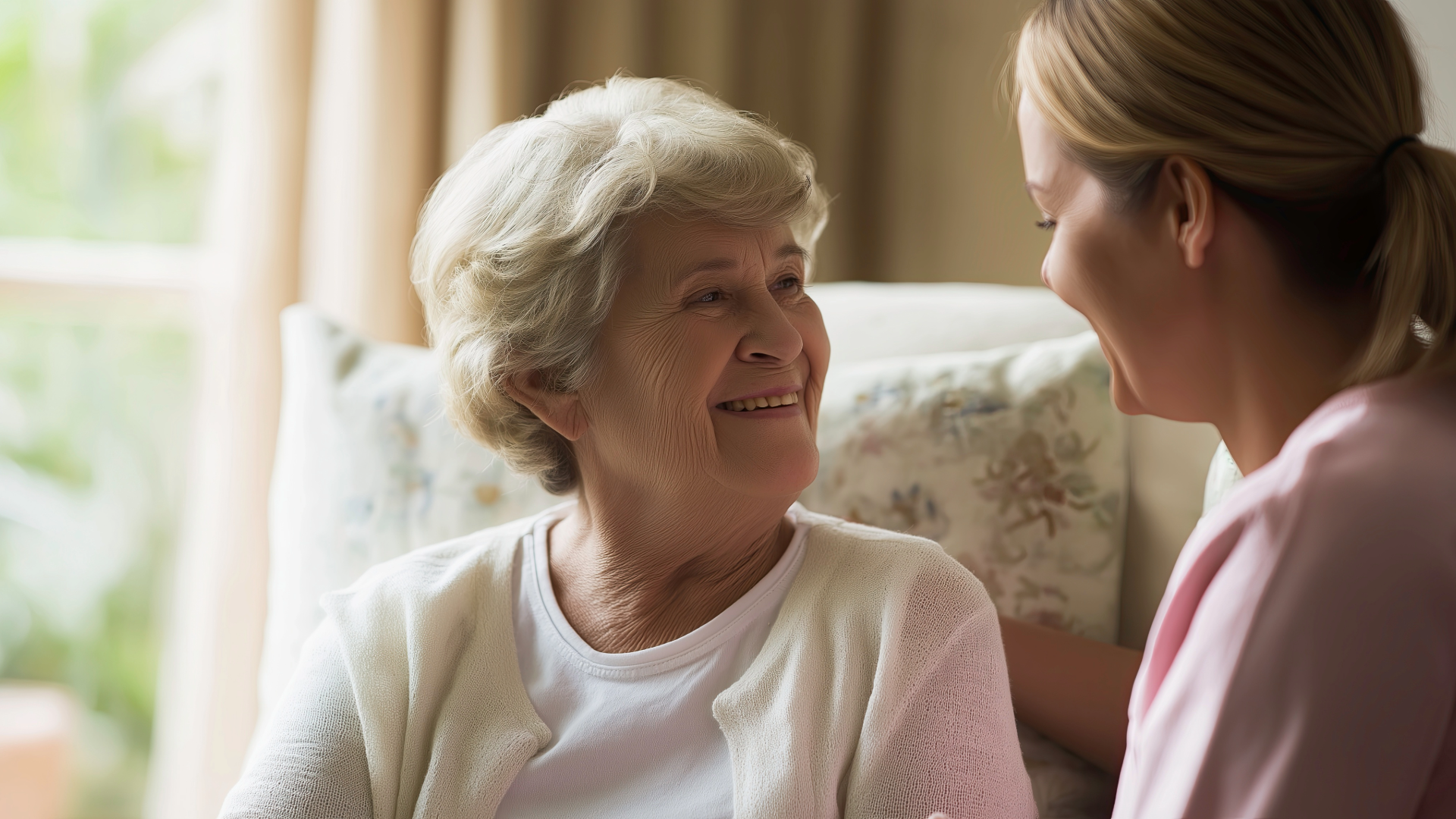 Woman smiles at caregiver in a light-filled room. The caregiver, with blonde hair, smiles back.