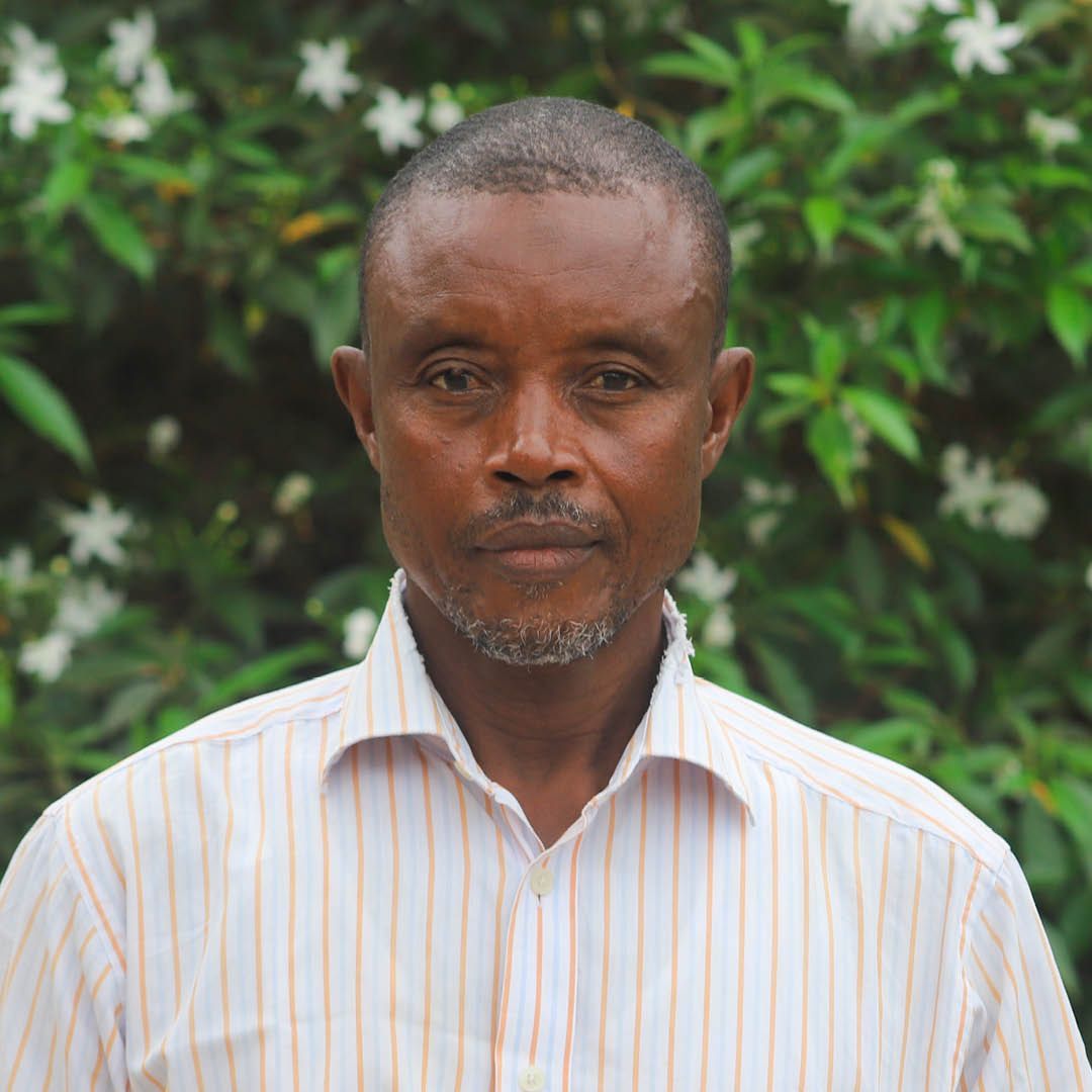 A man with a beard wearing a striped shirt is standing in front of a bush with white flowers.