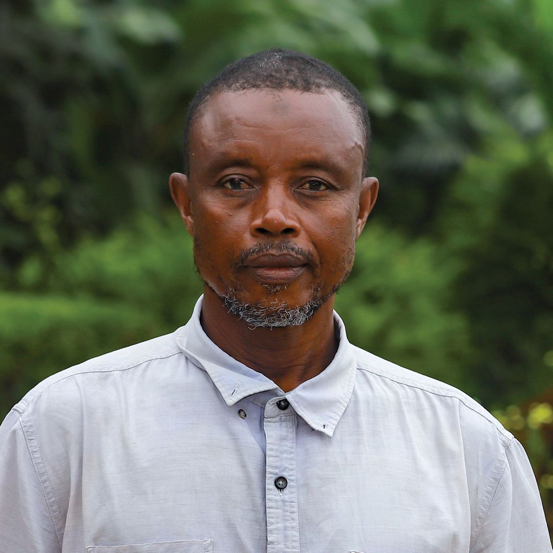 A man with a beard wearing a striped shirt is standing in front of a bush with white flowers.