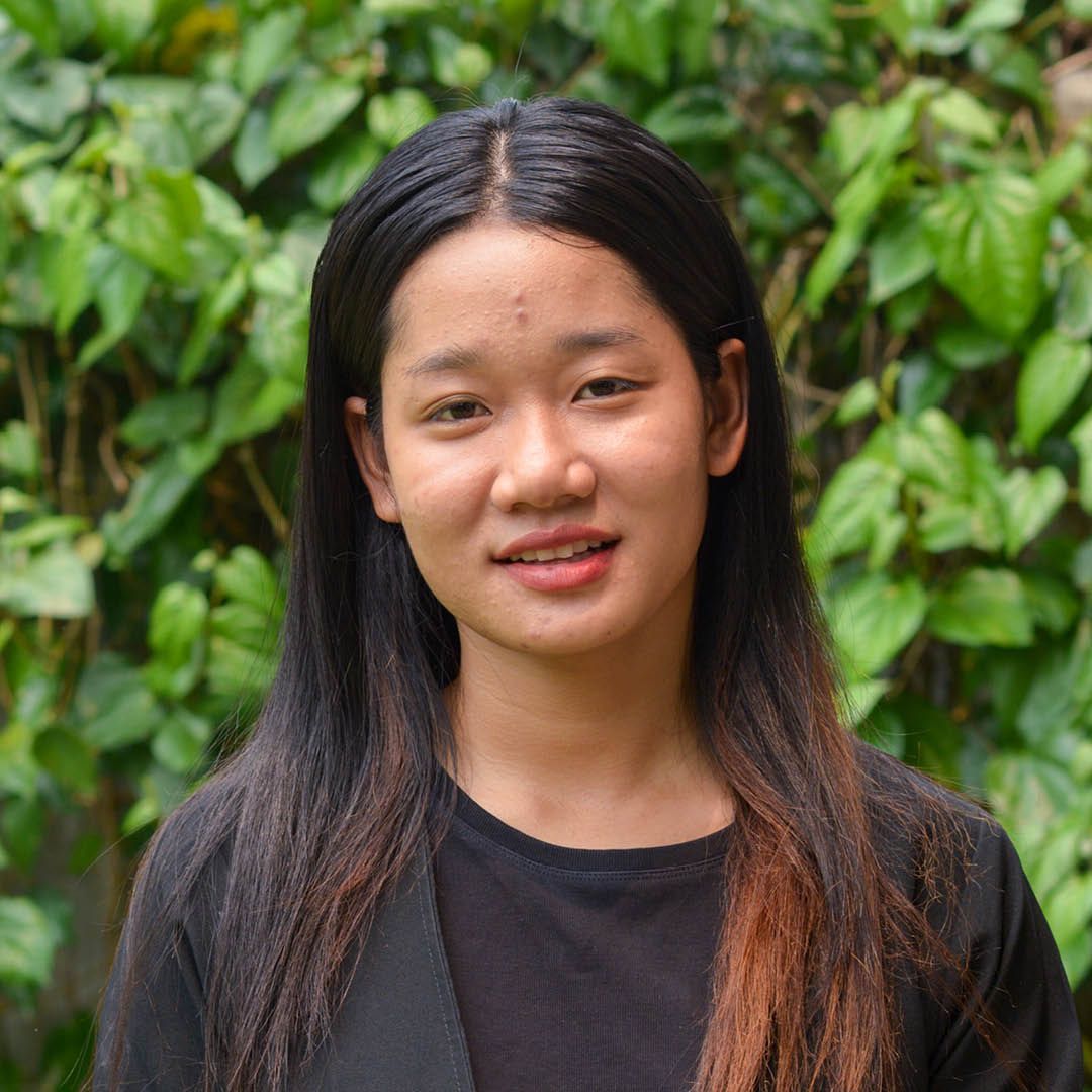 A young woman with long hair is standing in front of a bush.