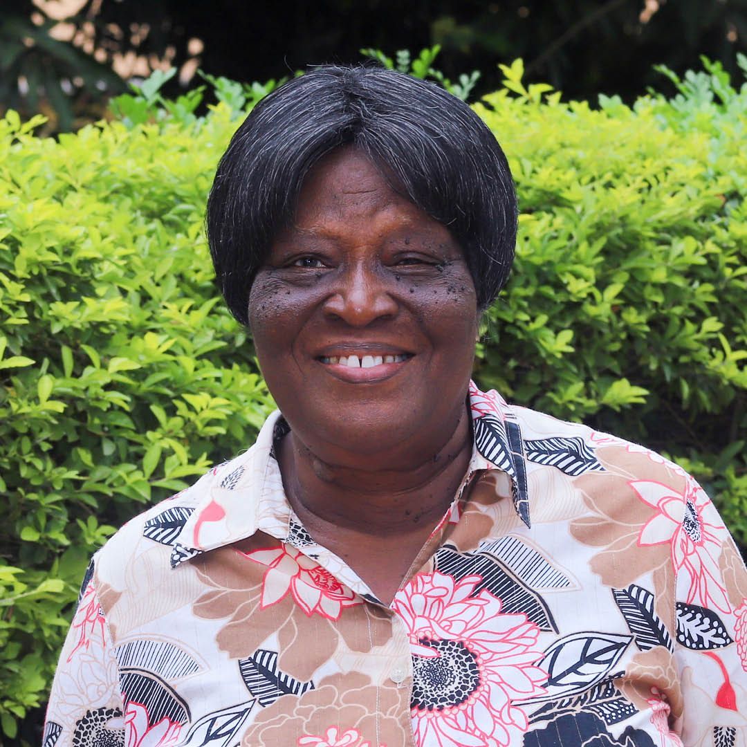 A woman in a floral shirt is smiling in front of a bush.