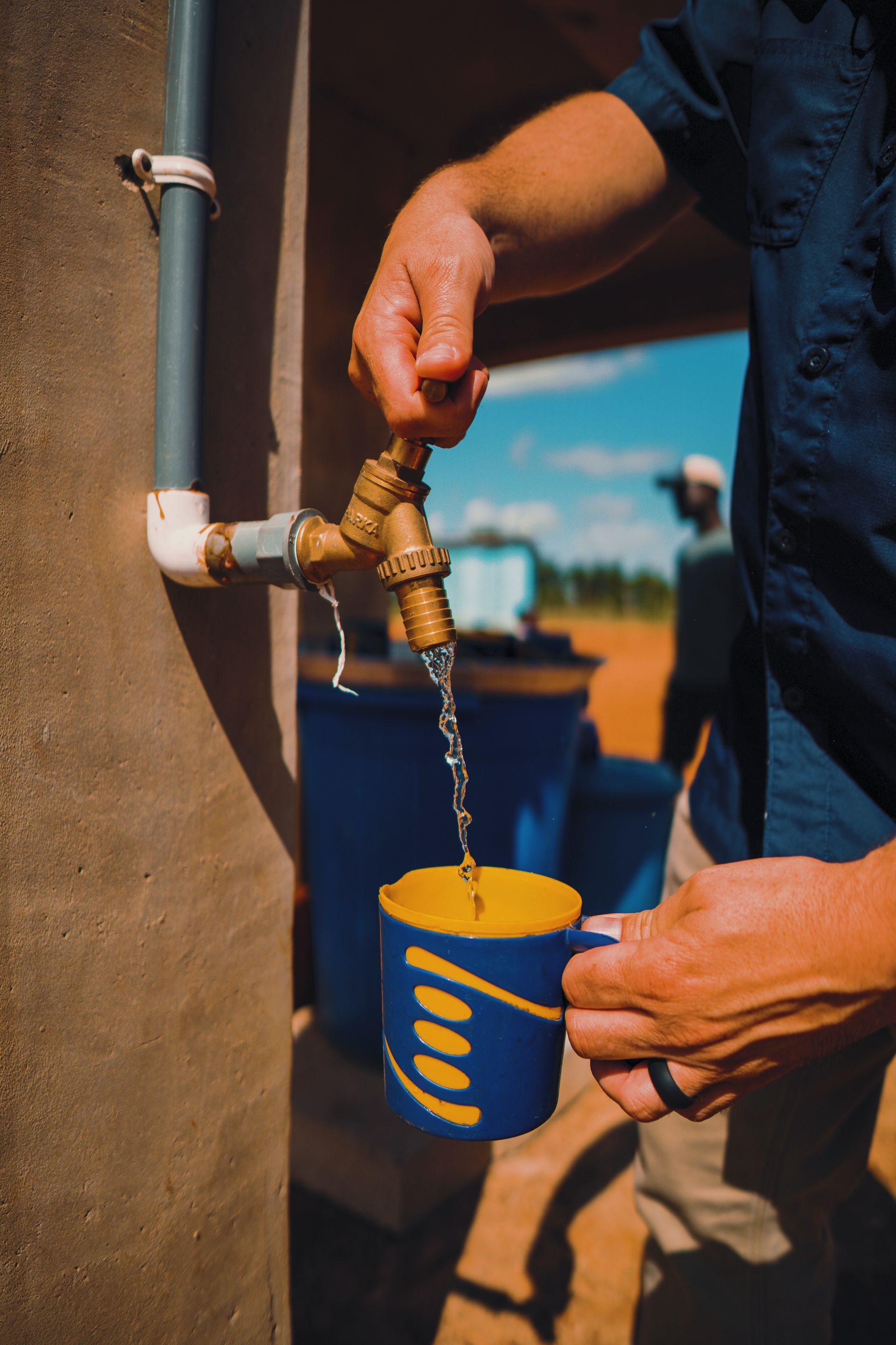 Person filling a blue and yellow mug with water from a tap outdoors.