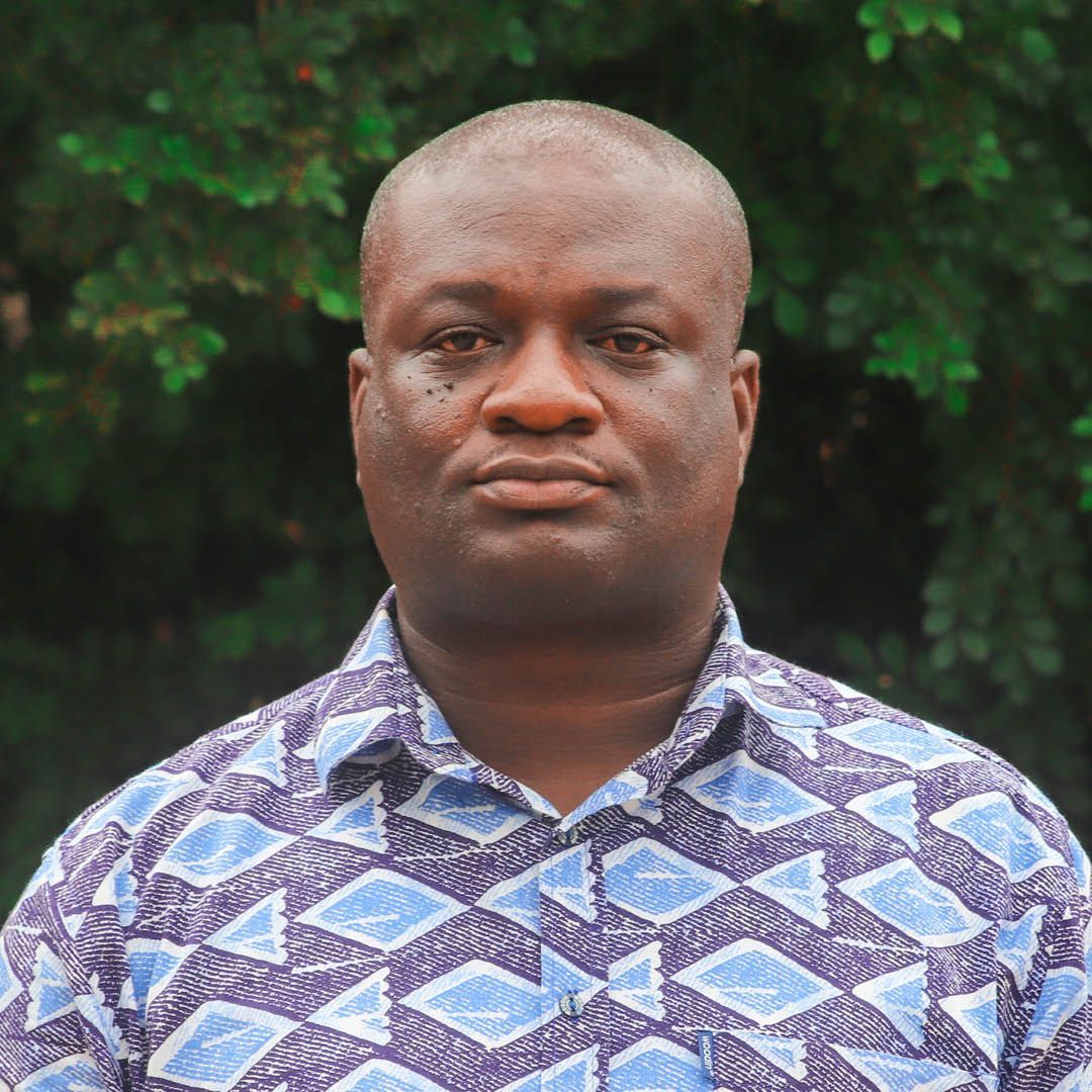 A man in a blue and purple shirt is standing in front of a tree.