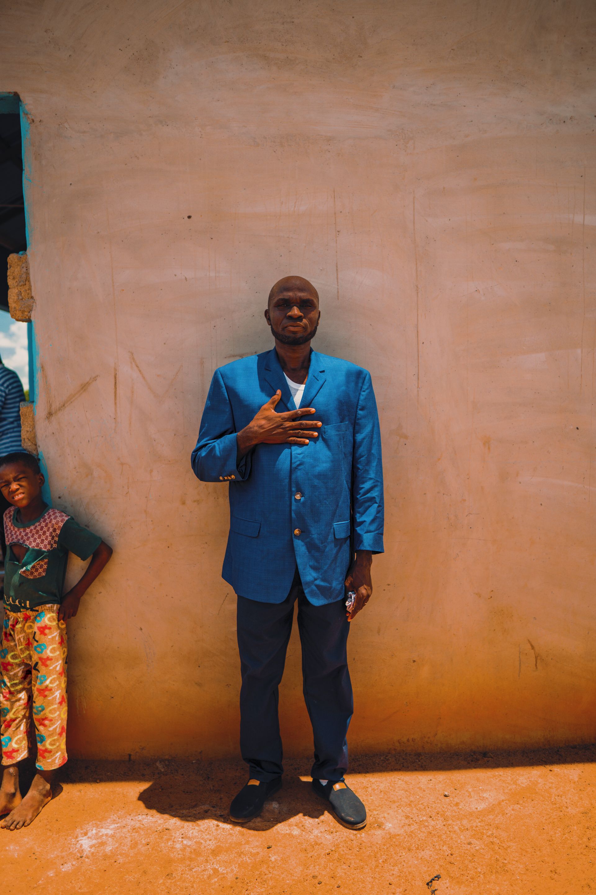 A man in a blue suit is standing in front of a wall with his hand on his chest.