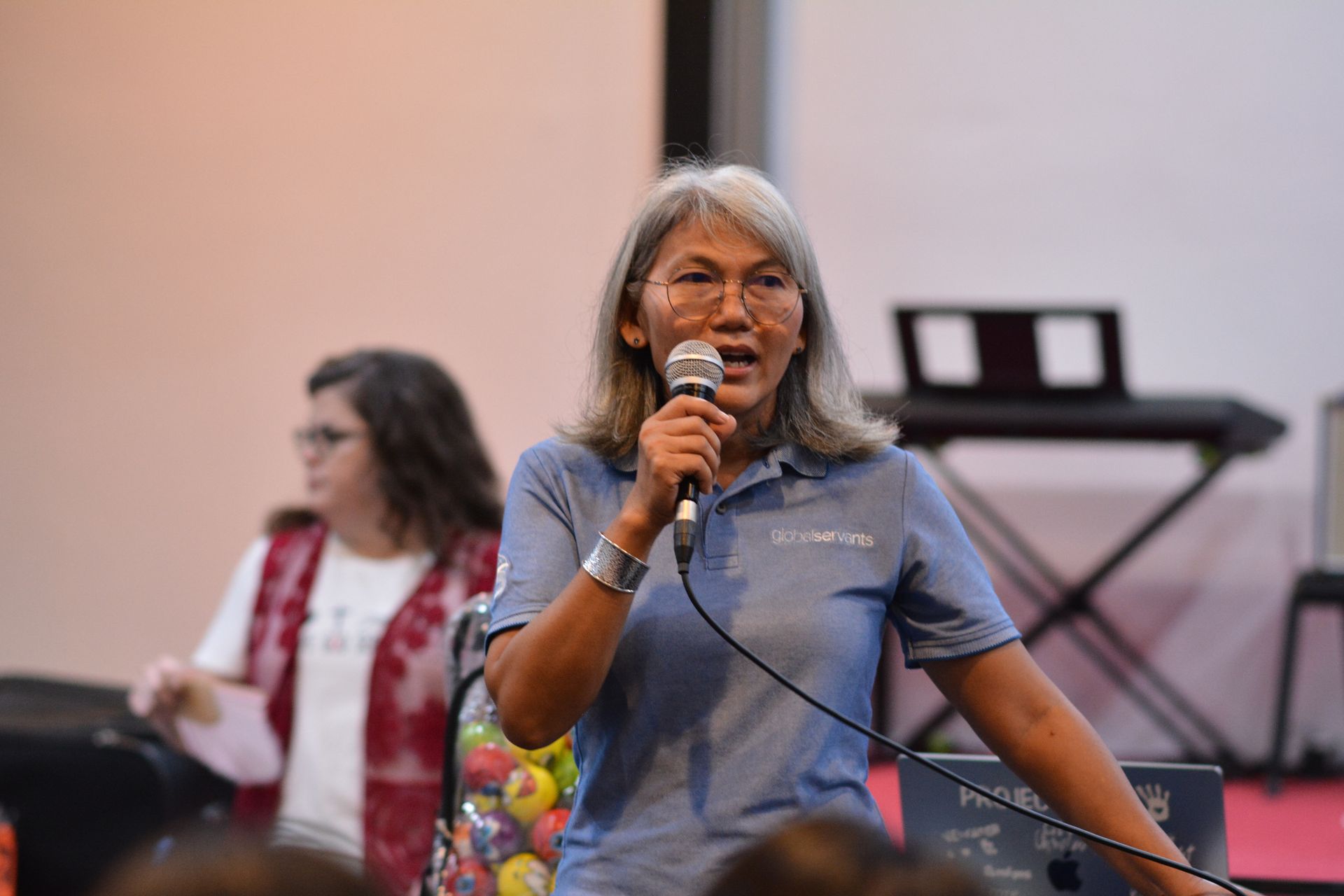 Woman speaking into microphone at an event, wearing a blue shirt. Another person is in the background.