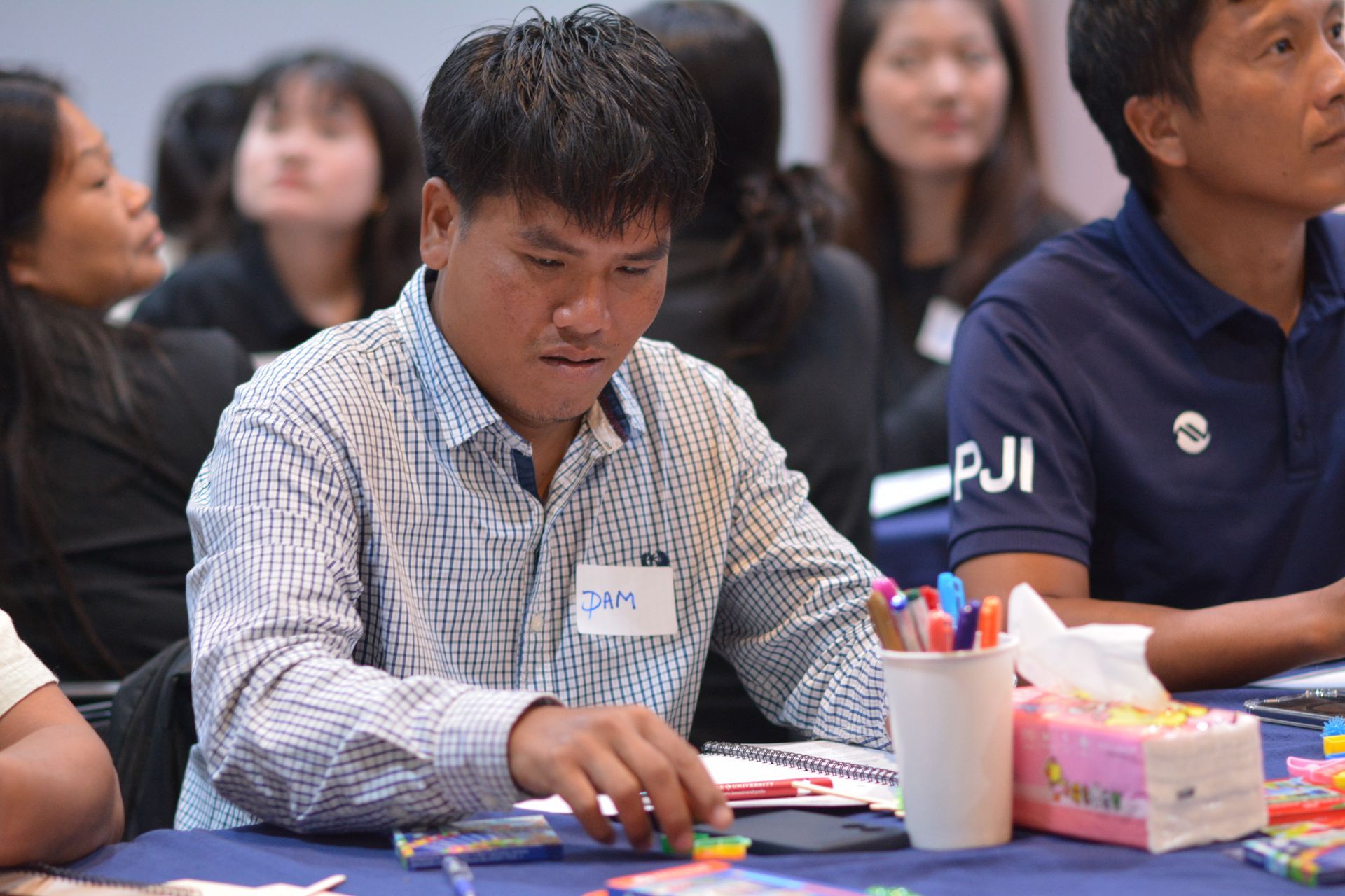 Man at table, wearing striped shirt, looking down at objects. Other people in the background.