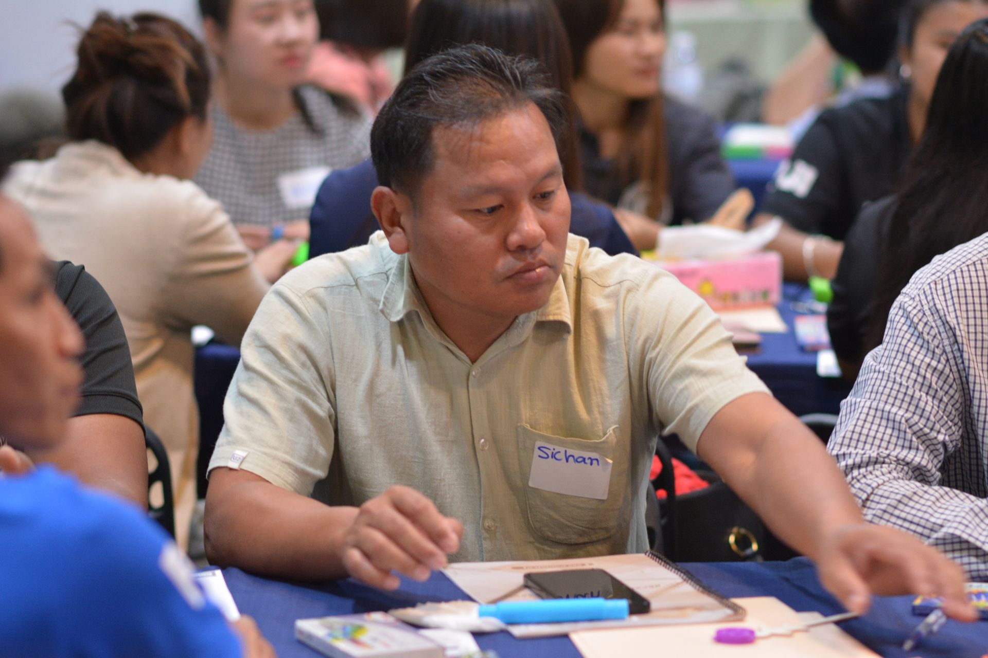 Man in light yellow shirt at a table with others, looking down, possibly participating in a meeting.