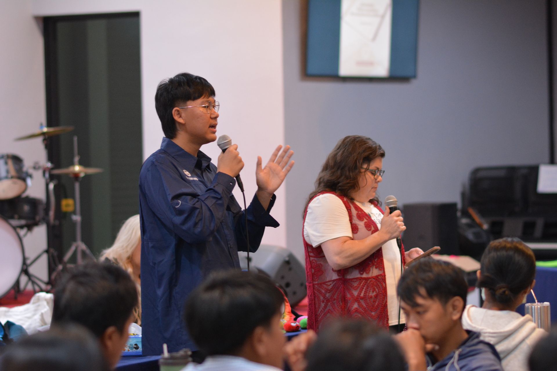 Two people with microphones addressing a group of people. One man in blue jacket, the woman in red vest.