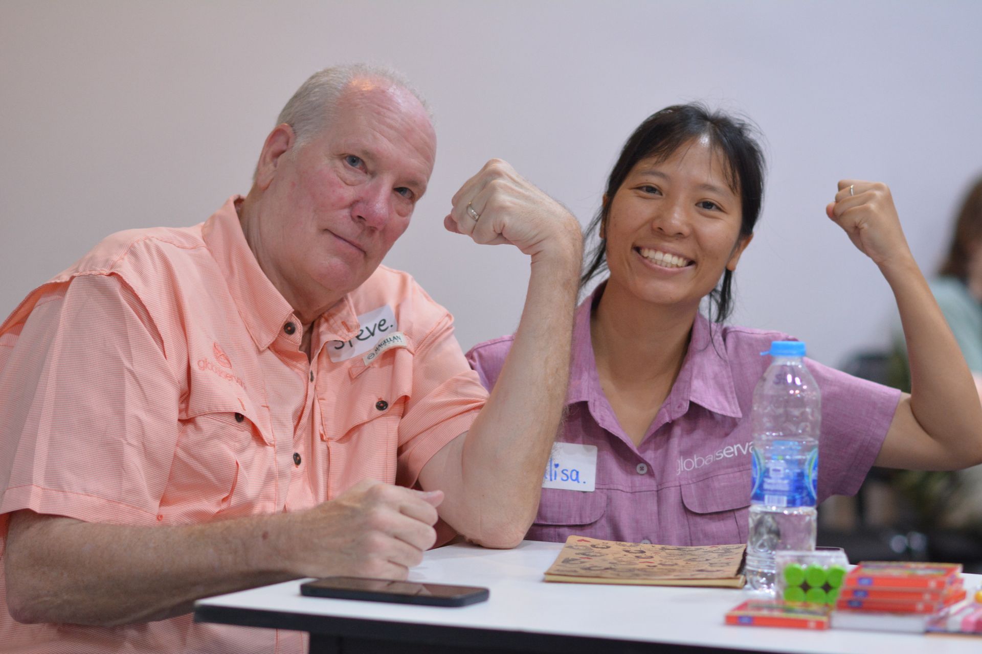 Man and woman smiling, flexing arms, sitting at table with water bottle.