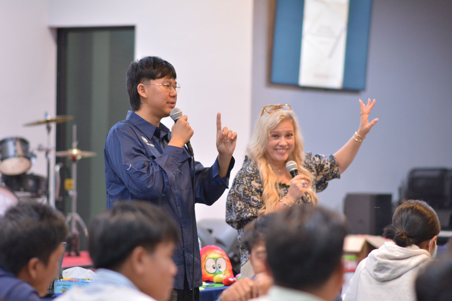 Man and woman speaking into microphones, gesturing to a seated audience in a room.