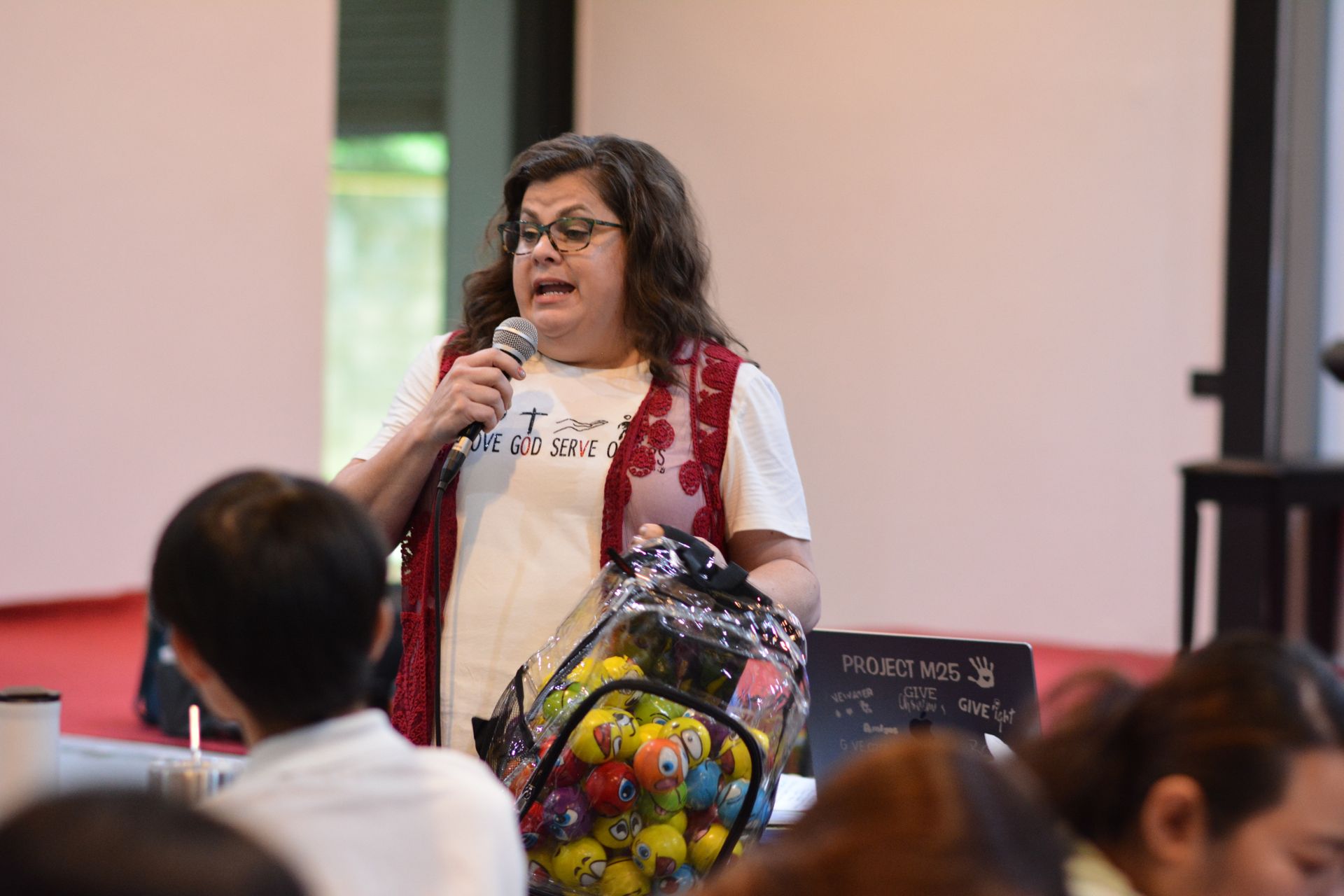 Woman speaking into a microphone, holding a bag of colorful balls; indoors at an event.