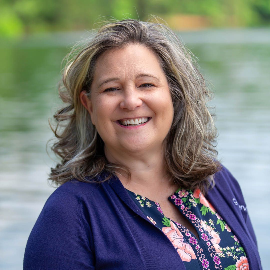 A woman in a purple sweater and floral shirt is smiling in front of a body of water.