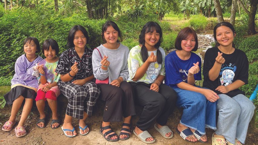 Seven smiling people making finger hearts, sitting outdoors by greenery.