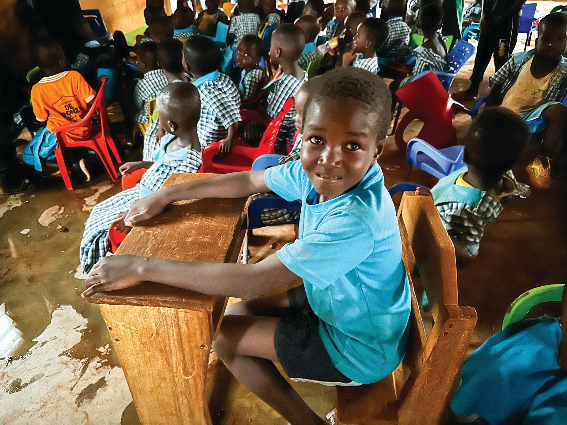 Young boy in blue shirt at a wooden desk smiles in a classroom with other children.