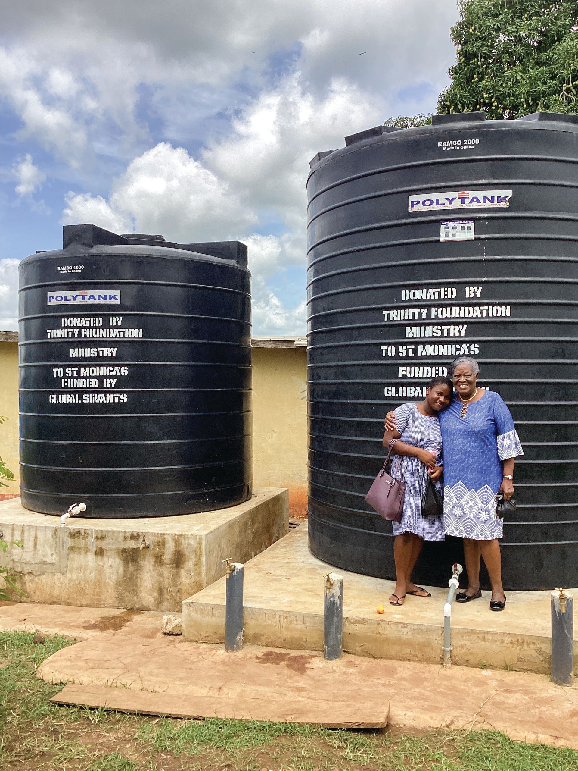 Two women are standing next to two large black water tanks.