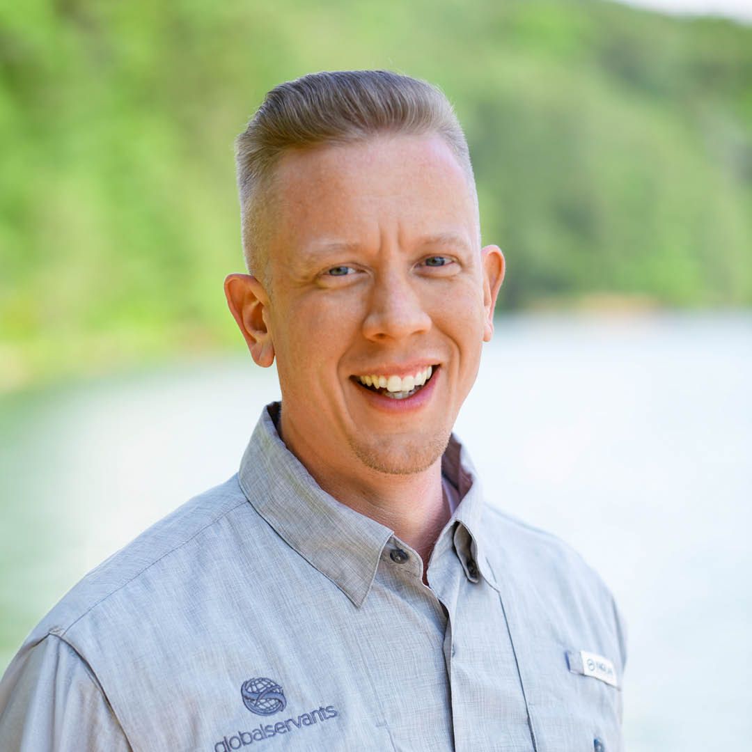 A man in a grey shirt is smiling in front of a body of water.