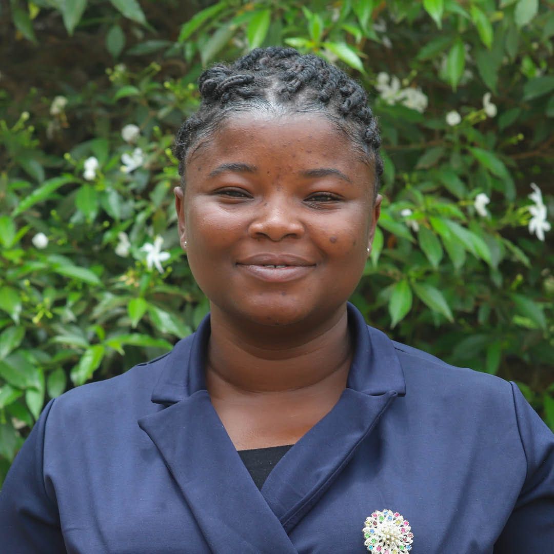 A woman in a blue jacket is smiling in front of a bush.