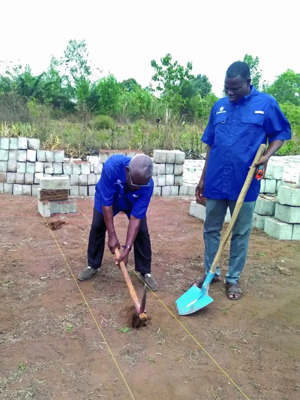 Two men in blue shirts are digging in the dirt with shovels