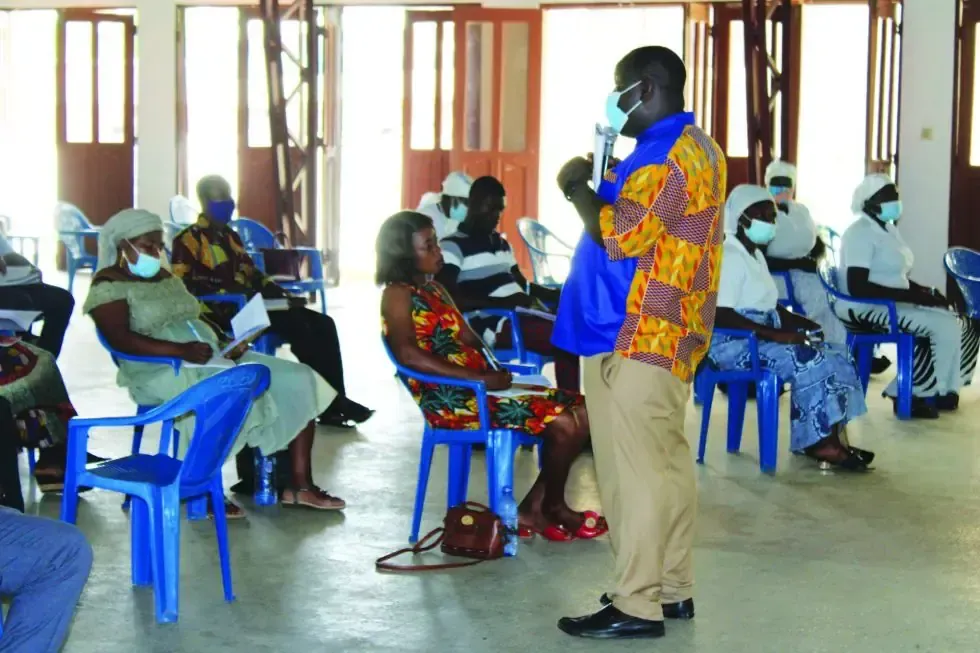 A man wearing a mask stands in front of a group of people sitting in chairs