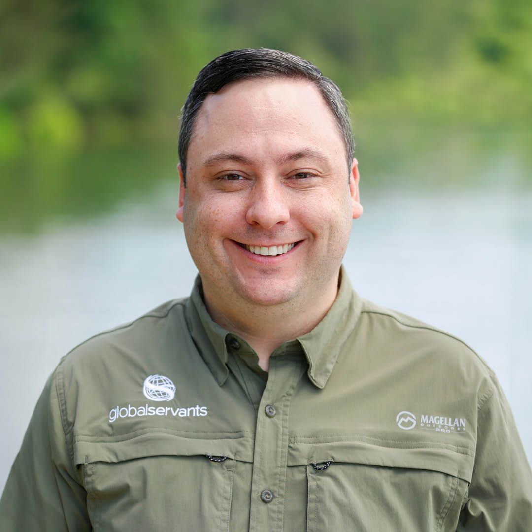 A man in a green shirt is smiling in front of a body of water.