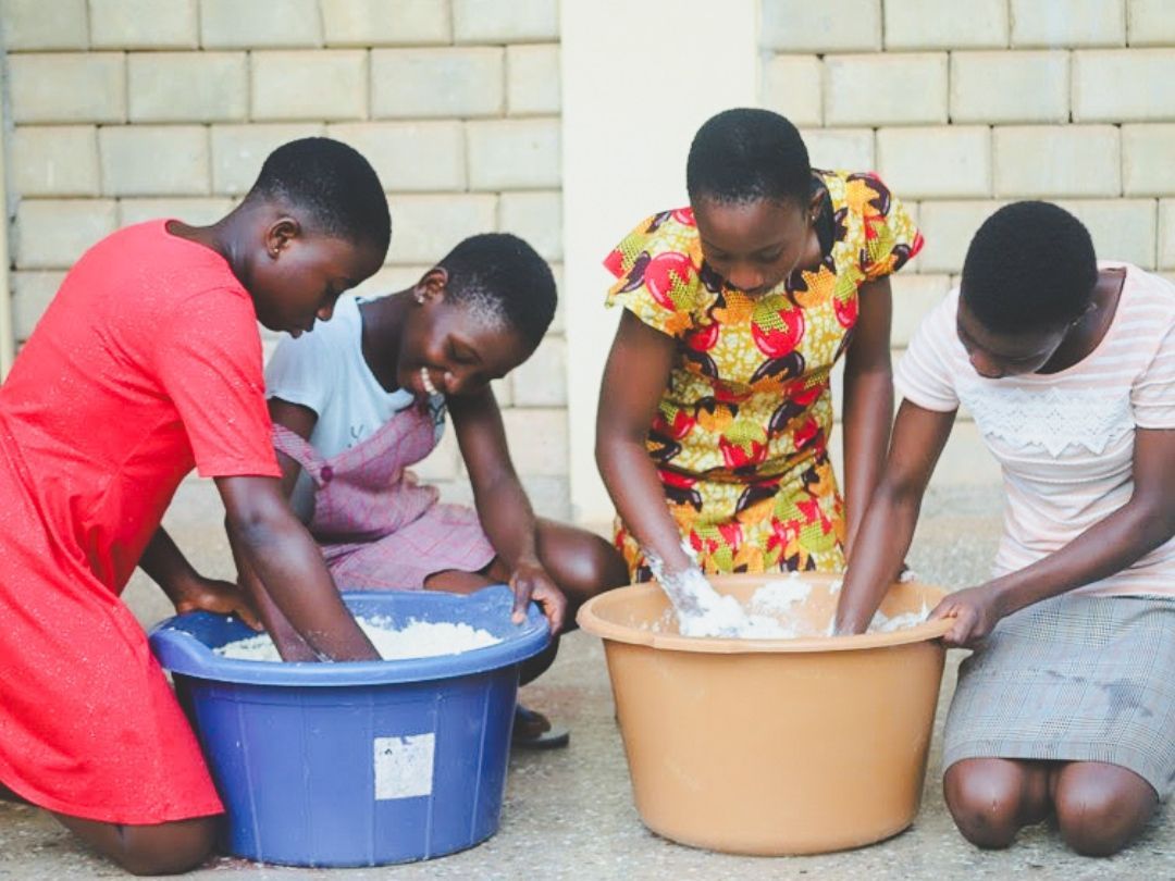 Four people mixing ingredients in tubs on the ground, likely cooking outdoors.