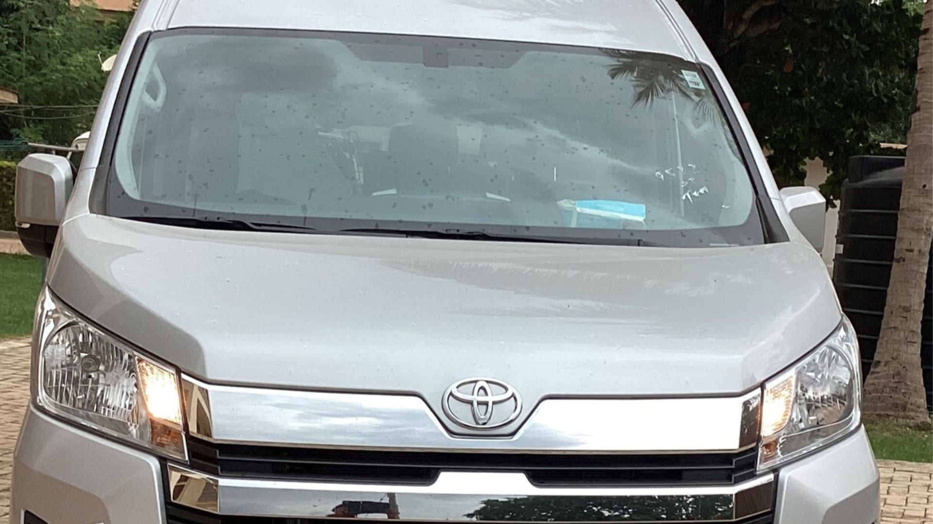 Silver Toyota Hiace van parked outdoors, headlights on.