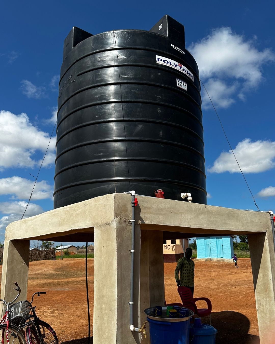 Black water tank on a concrete stand; person stands nearby. Blue sky, outdoor setting.
