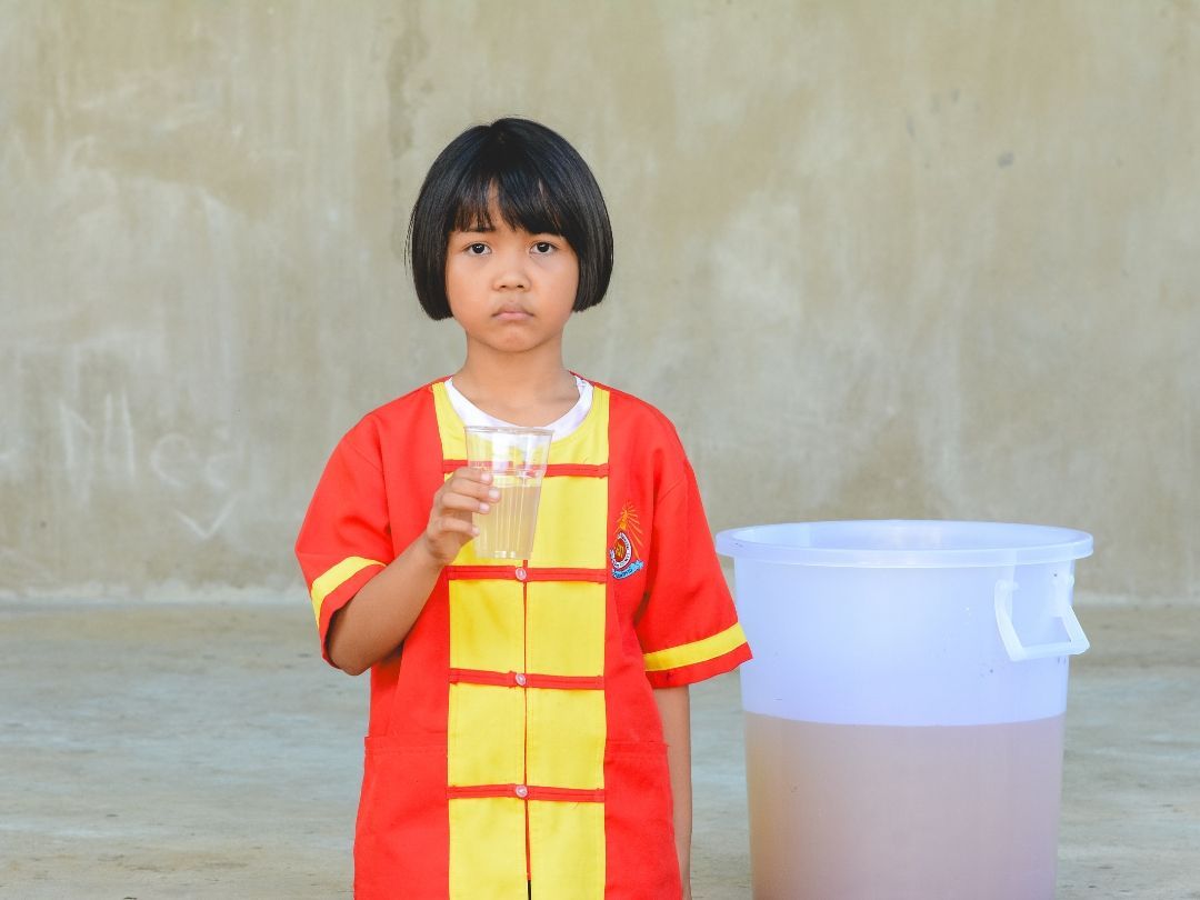 Girl in red and yellow outfit holding a glass, standing next to a large container.