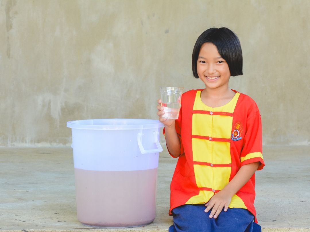 Girl smiling, holding glass of water, beside a water container. She's wearing red and yellow shirt.