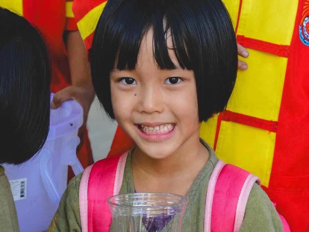 Girl with black hair and a pink backpack smiles, holding a clear glass. Bright red and yellow background.