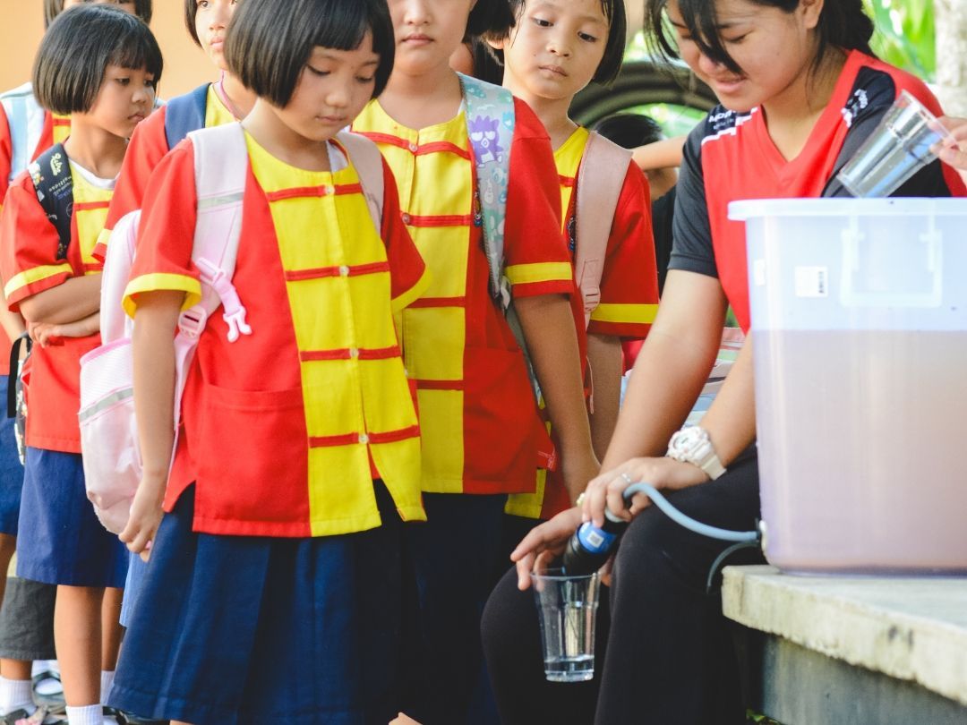 Schoolchildren in red and yellow uniforms lining up for water from a container.
