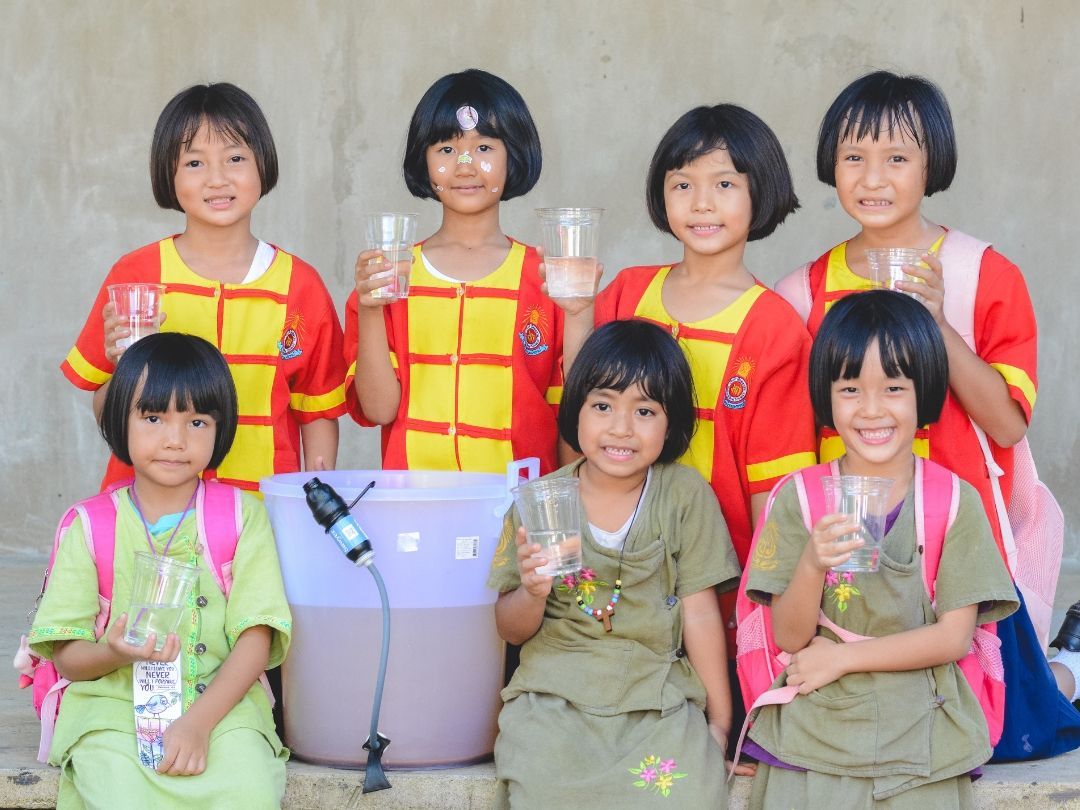 Seven children smiling, holding glasses, near a water filter. They wear colorful clothes outside.