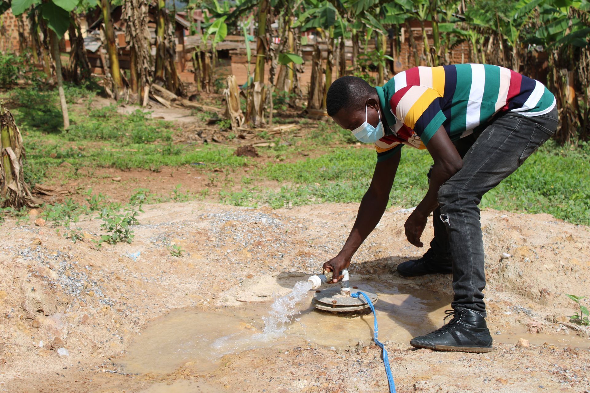 A man wearing a mask is washing his hands with a hose.