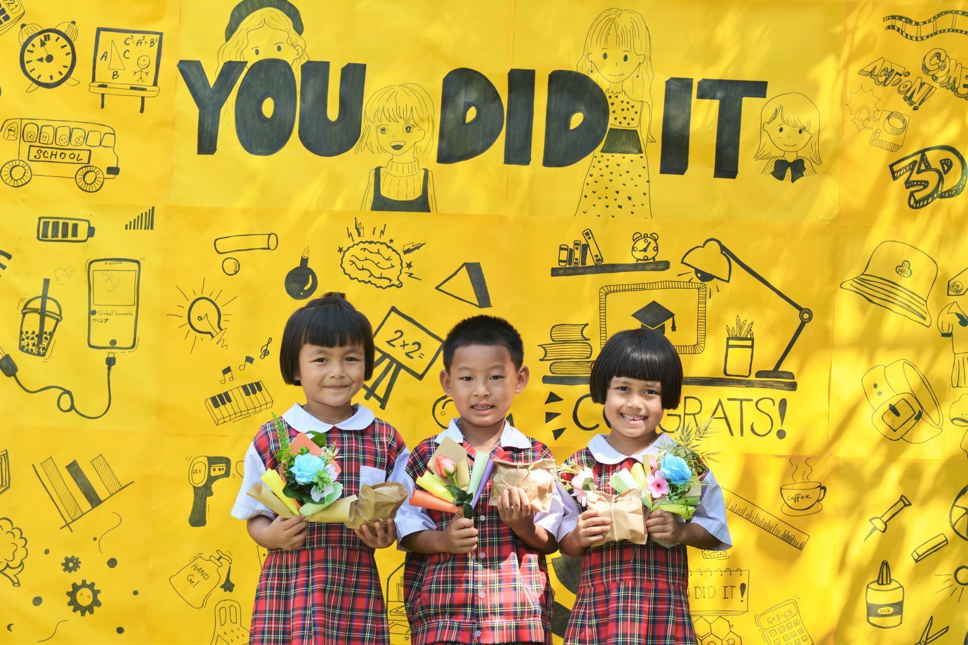 Three children are standing in front of a yellow sign that says you did it.