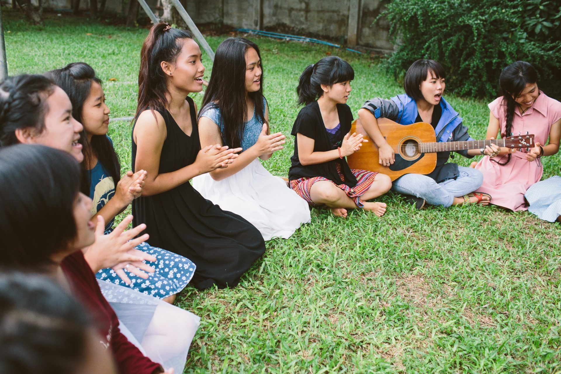 A group of young women are sitting on the grass singing and playing guitars.