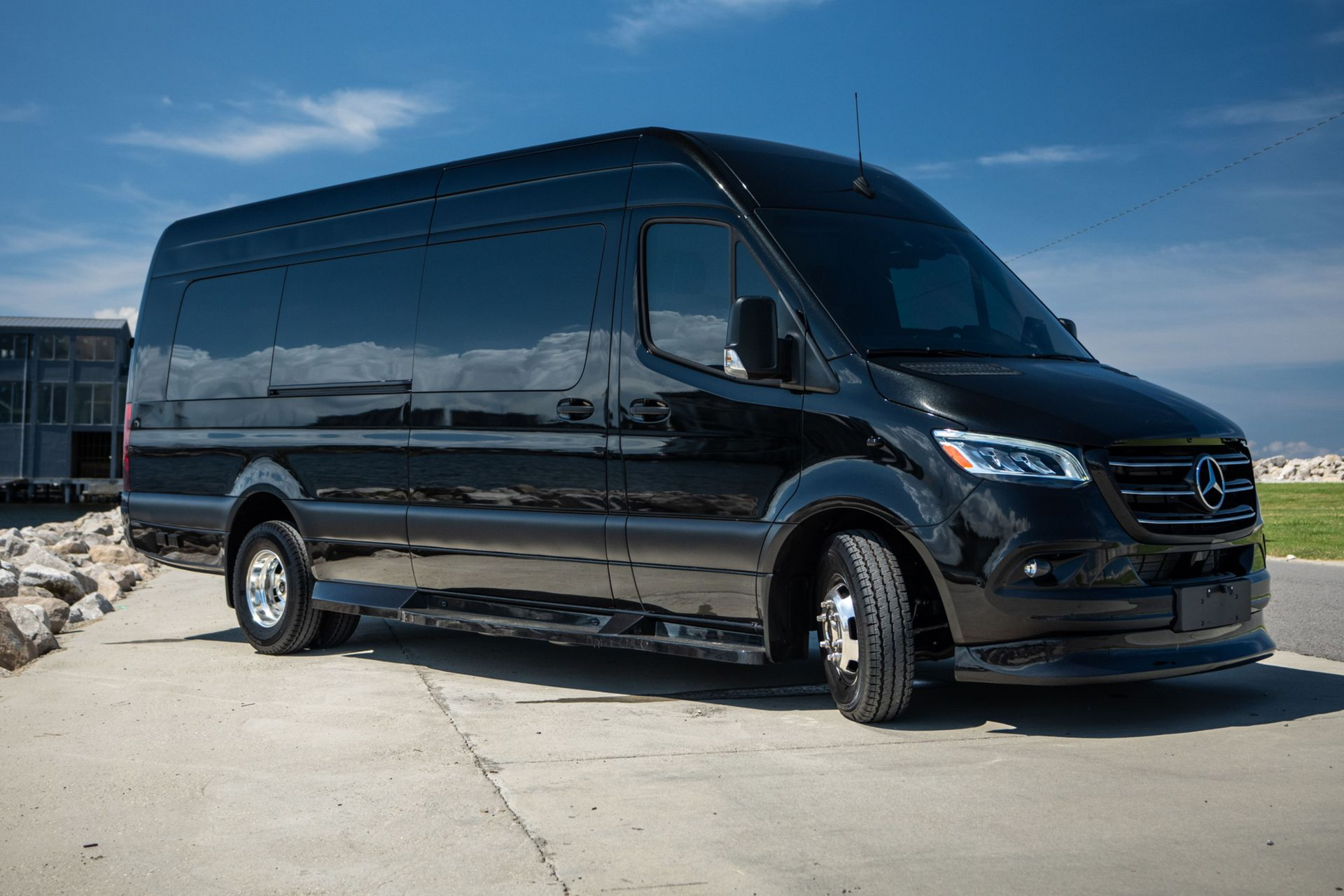 A black Mercedes-Benz Sprinter van parked on an asphalt lot near a building by the water under a clear blue sky.