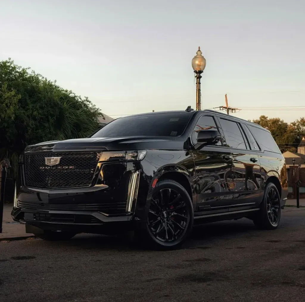 A black Cadillac Escalade SUV parked on a paved lot at sunset with a streetlamp in the background.