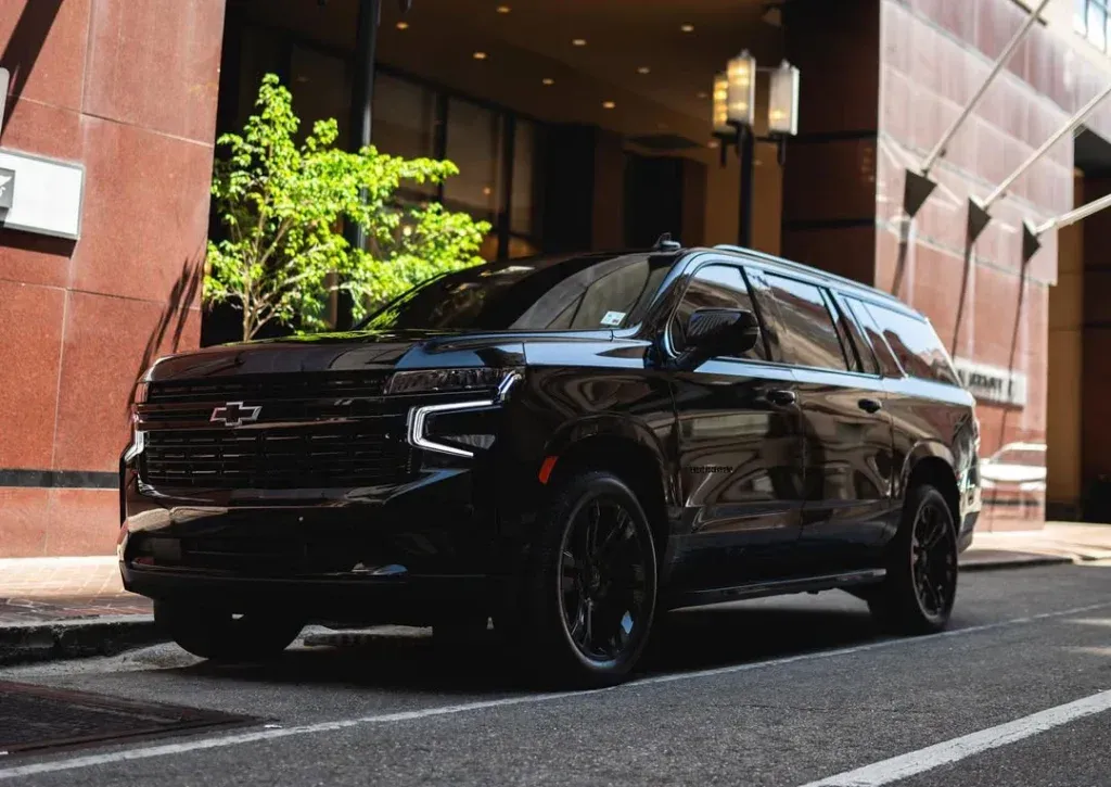 A black Chevrolet Suburban parked on a city street next to a modern building with a tree in the background.