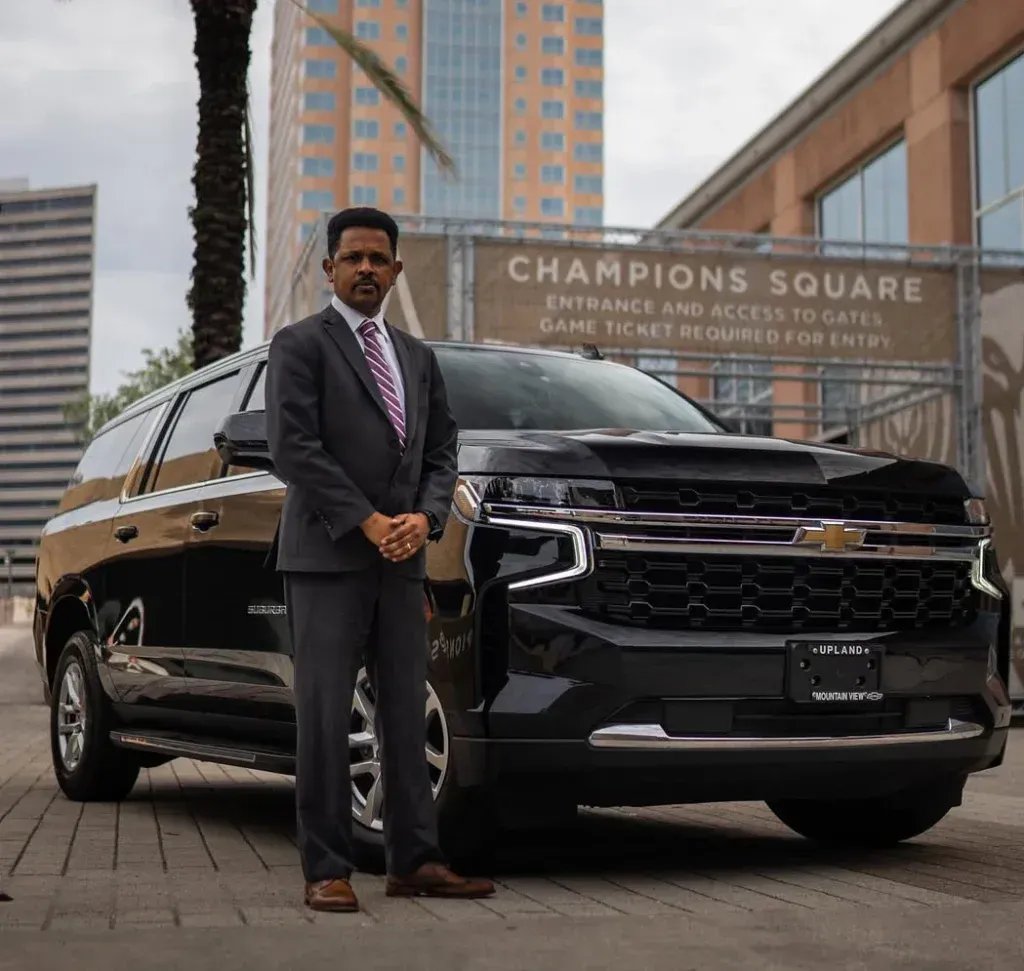 A person in a suit stands beside a black SUV parked in front of Champions Square.
