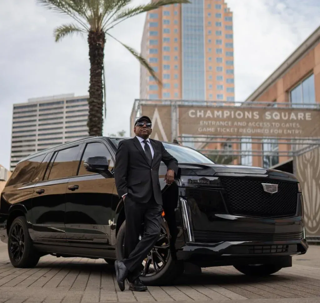 A person in a suit poses beside a black Cadillac SUV in front of Champions Square in an urban setting.