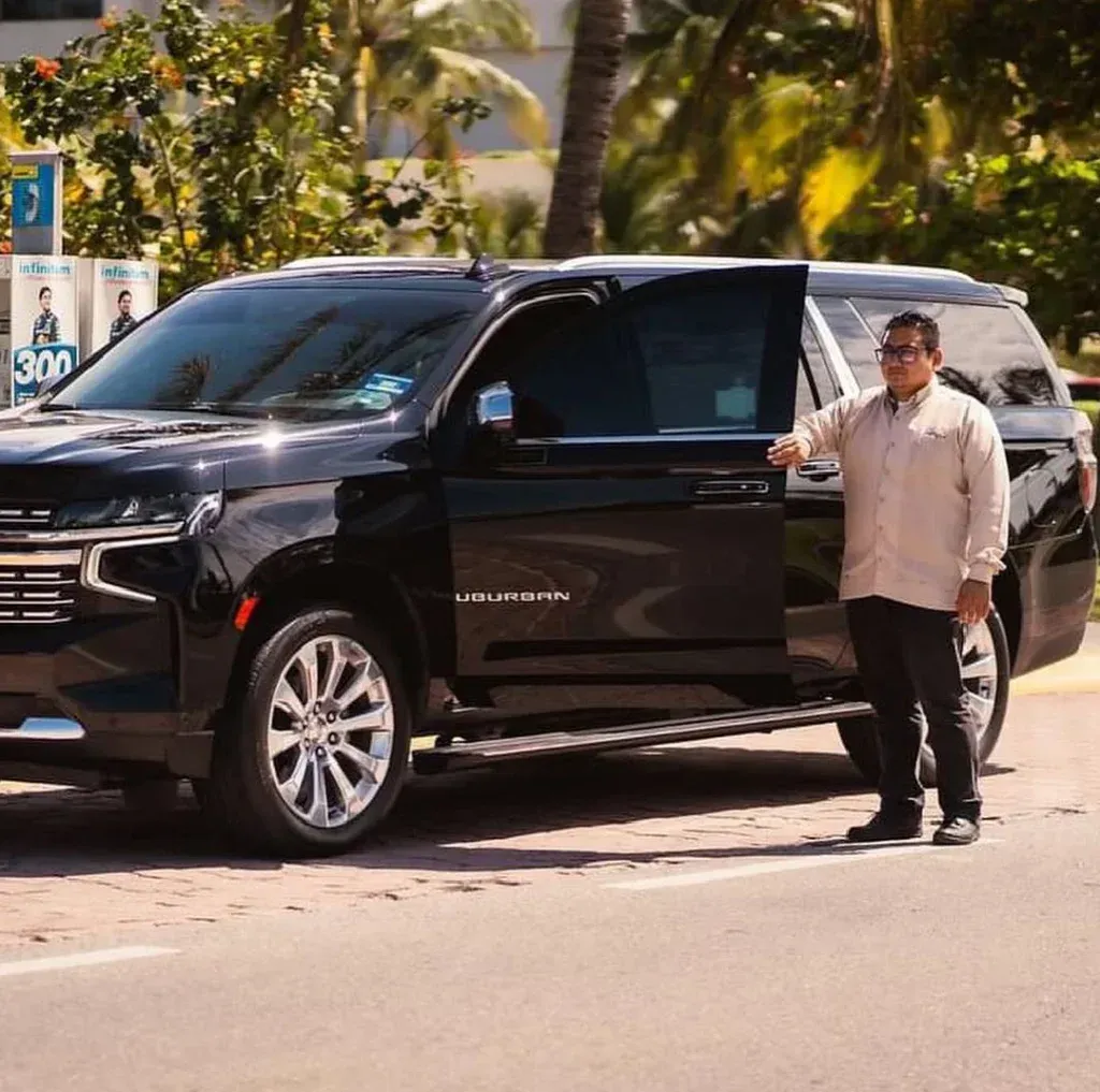 A person in a light shirt and dark trousers stands by the open door of a black SUV parked on a paved road.