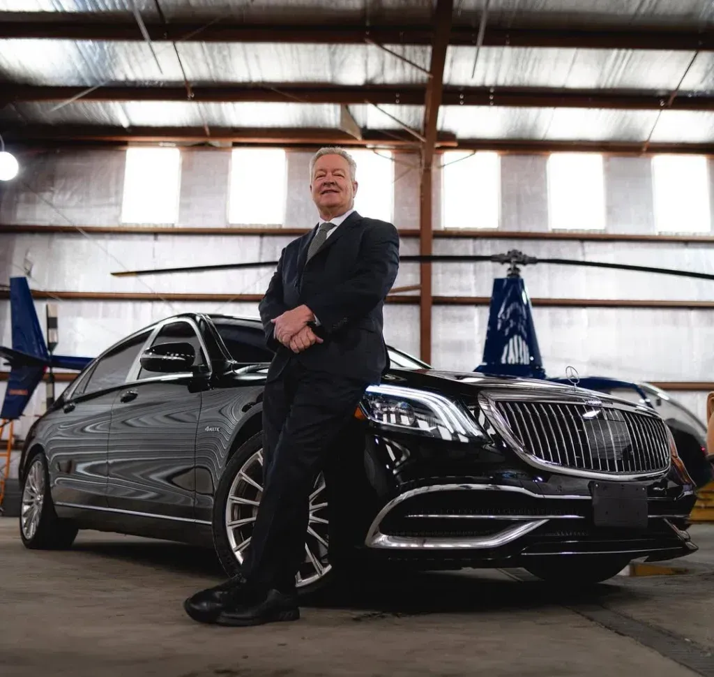A person in a suit leans against a black luxury car inside a hangar with two helicopters in the background.