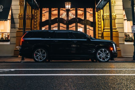 A dark SUV parked on a city street in front of a brick building with many rectangular windows.