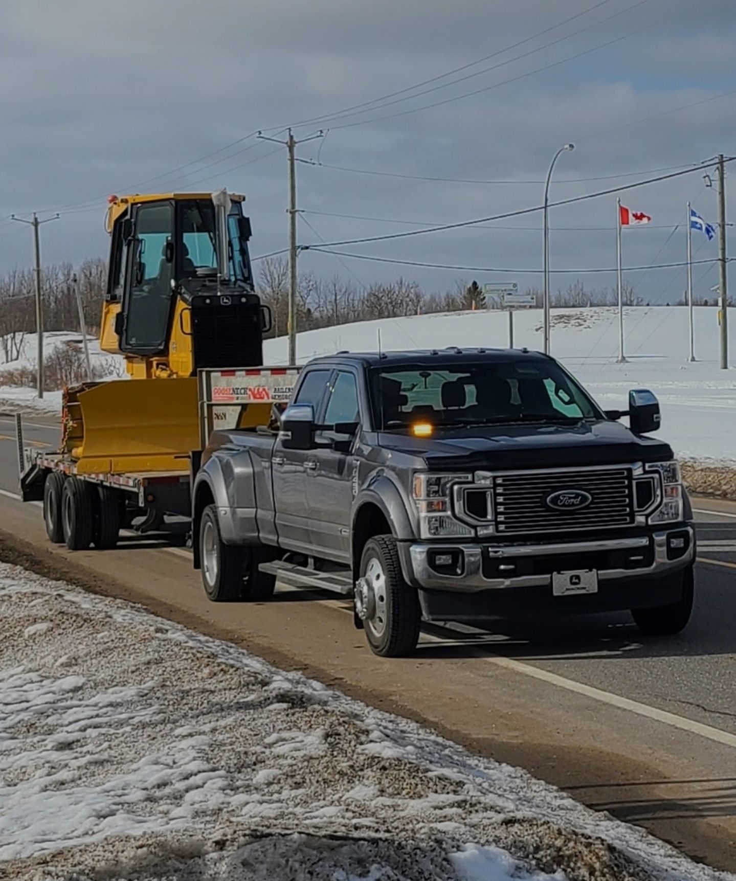 Une camionnette noire tracte une remorque jaune d'excavatrice sur le bord d'une route enneigée, près de drapeaux et de poteaux électriques.