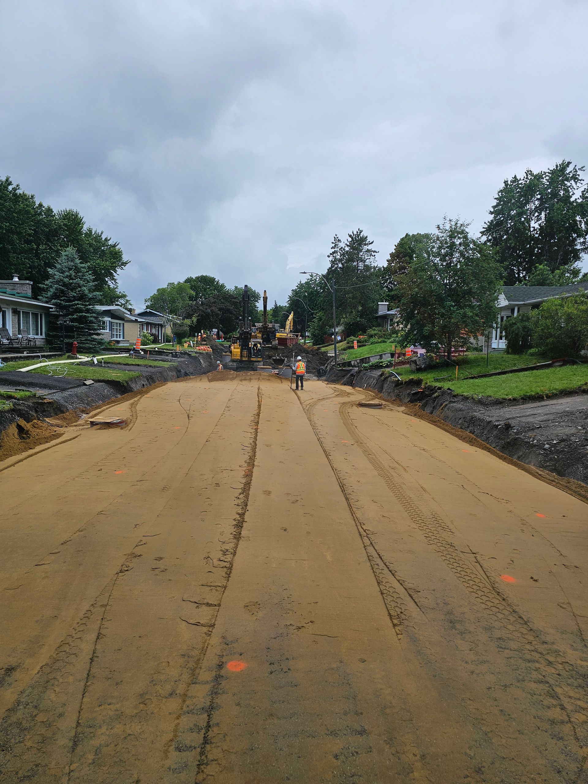 Large route boueuse en travaux, avec des ouvriers et du matériel au loin, des arbres et des maisons de part et d'autre.