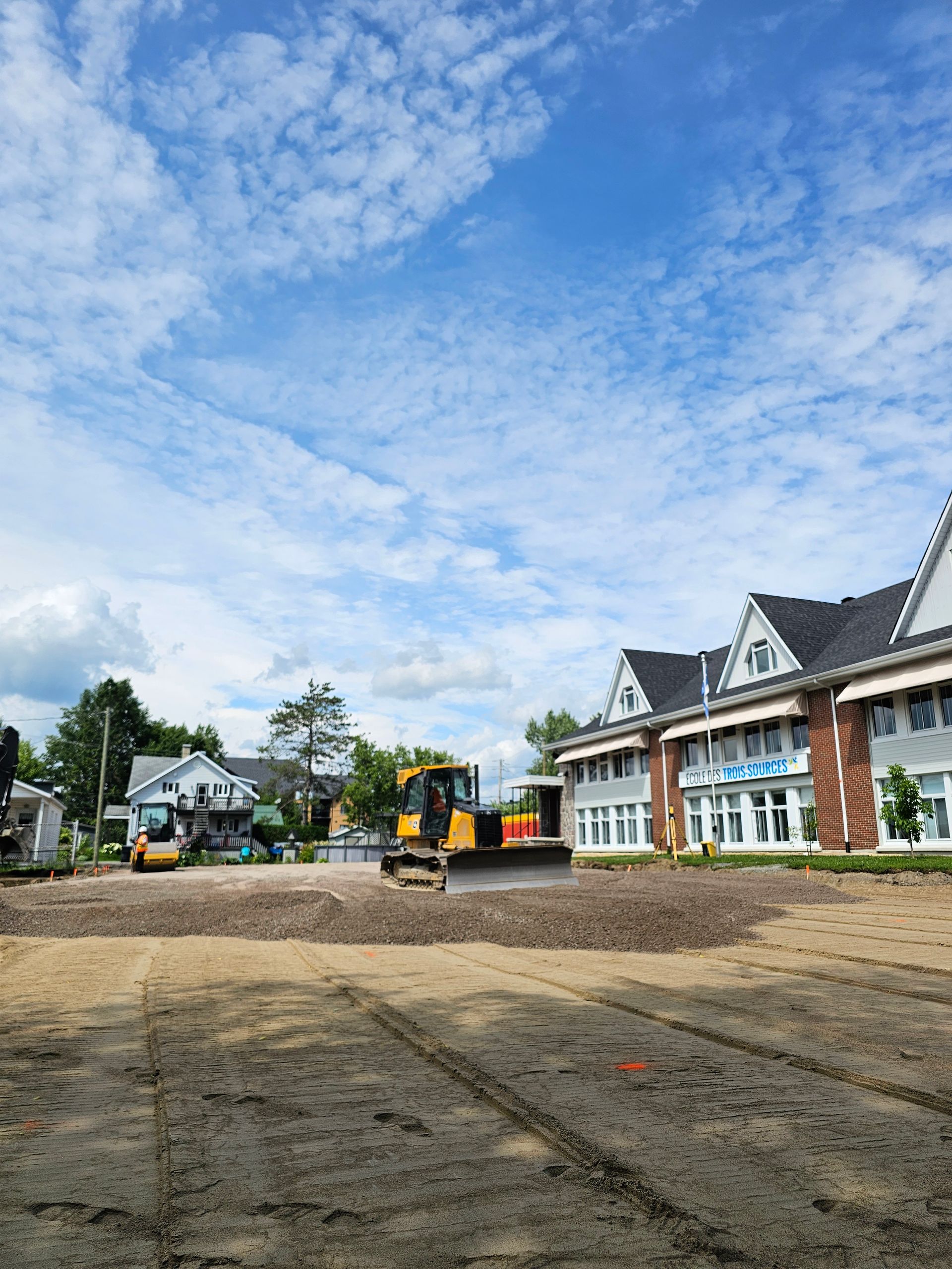 Chantier de construction avec une excavatrice à côté d'un bâtiment en briques sous un ciel bleu partiellement nuageux