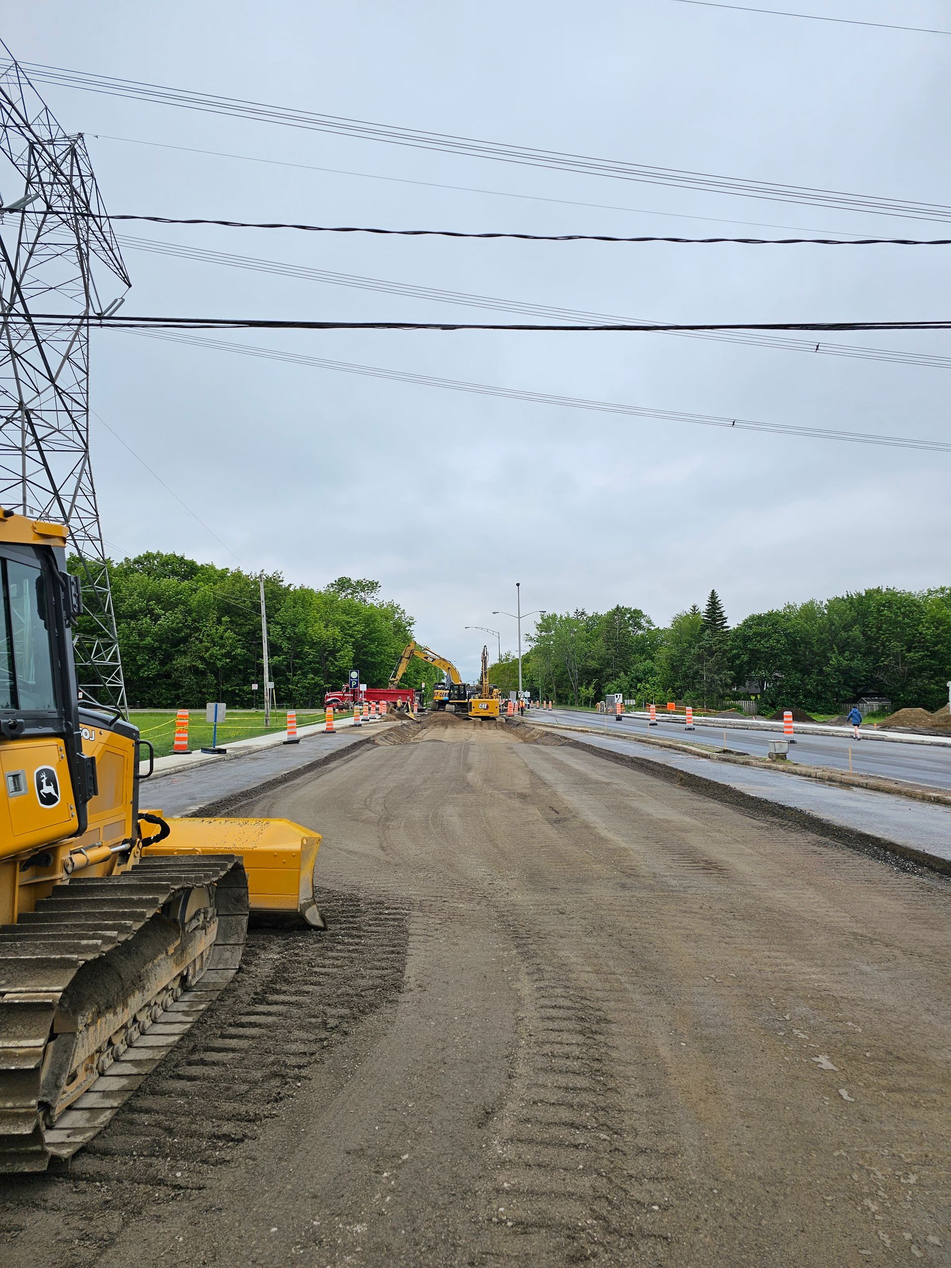 Chantier routier avec une chaussée en gravier, une excavatrice, des lignes électriques et des ouvriers au loin.